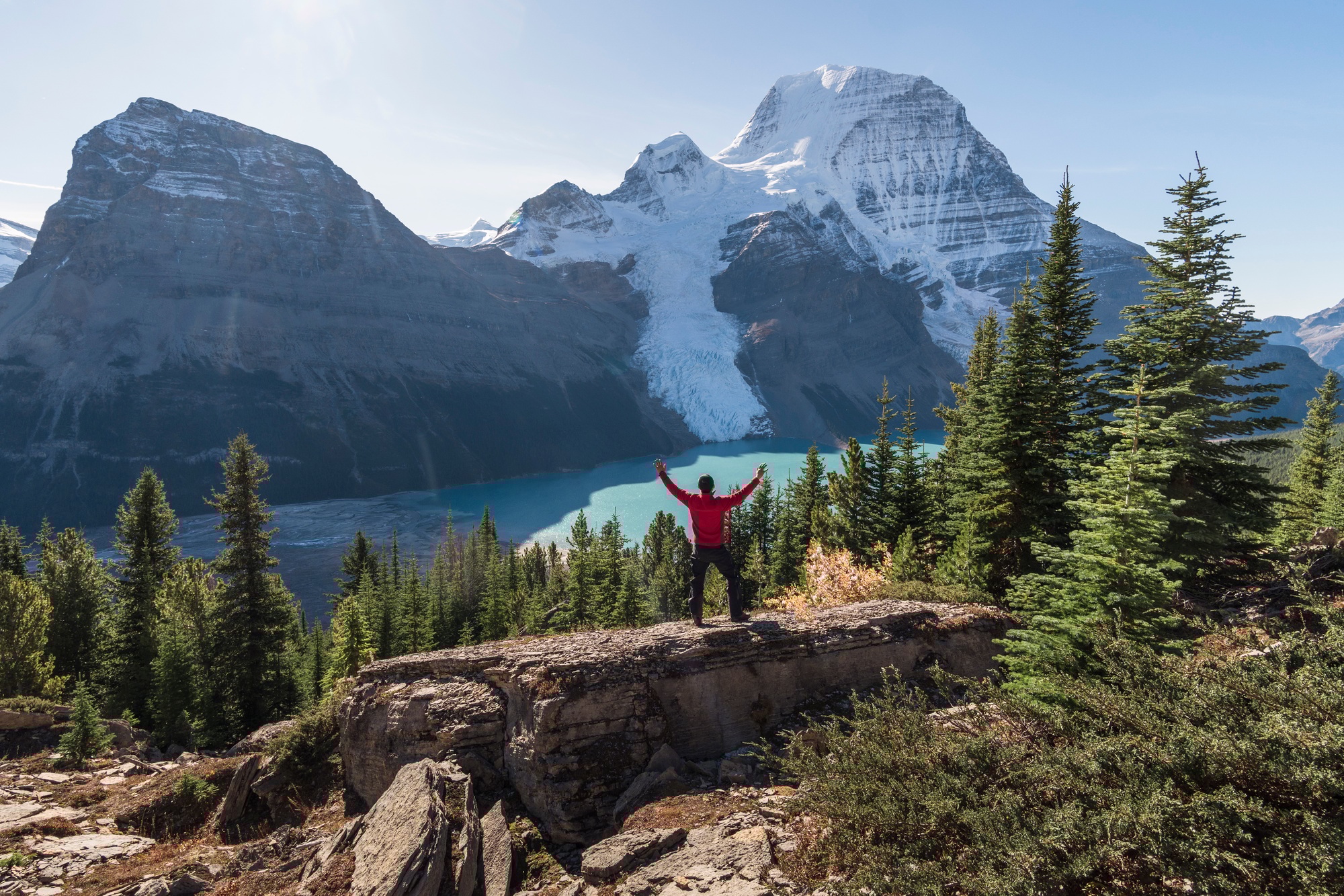 Man with arms raised, on a ridge above Berg lake and mountains.